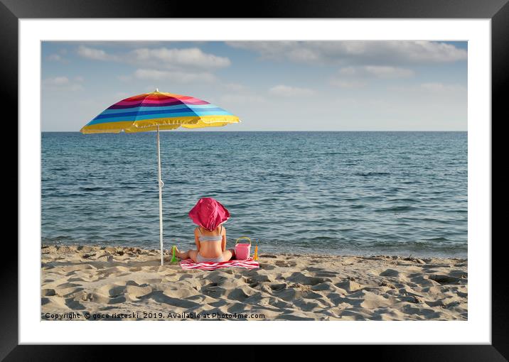 little girl with hat sitting under sunshade on bea Framed Mounted Print by goce risteski