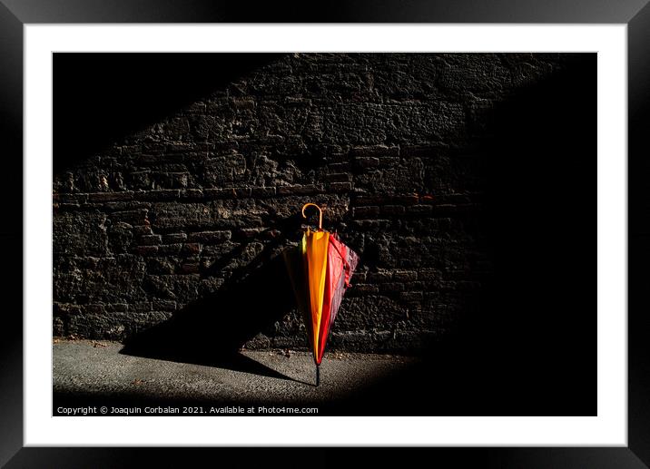 A beautiful colored umbrella rests folded against a brick wall i Framed Mounted Print by Joaquin Corbalan