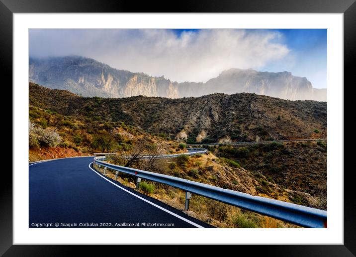 Winding road leads to high mountains with clouds to escape the r Framed Mounted Print by Joaquin Corbalan