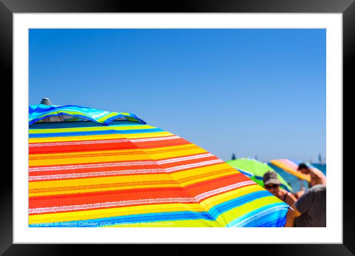 Colorful beach umbrella stuck in the sand surrounded by a group of bathers in summer, near the Mediterranean sea. Framed Mounted Print by Joaquin Corbalan