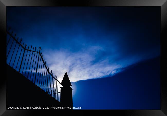 Silhouette against the sun of a high wall and metal fence with an intense blue sky in the background. Framed Print by Joaquin Corbalan