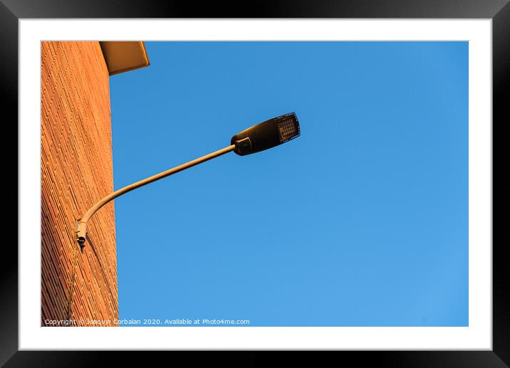Lamppost on the red wall of a building, by day, with a blue sky background. Framed Mounted Print by Joaquin Corbalan