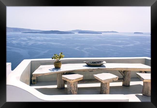 Santorini, Greece: A pot with flower or plant and a plate on a wooden table with wooden chair against beautiful sea ocean background with mountains Framed Print by Arpan Bhatia