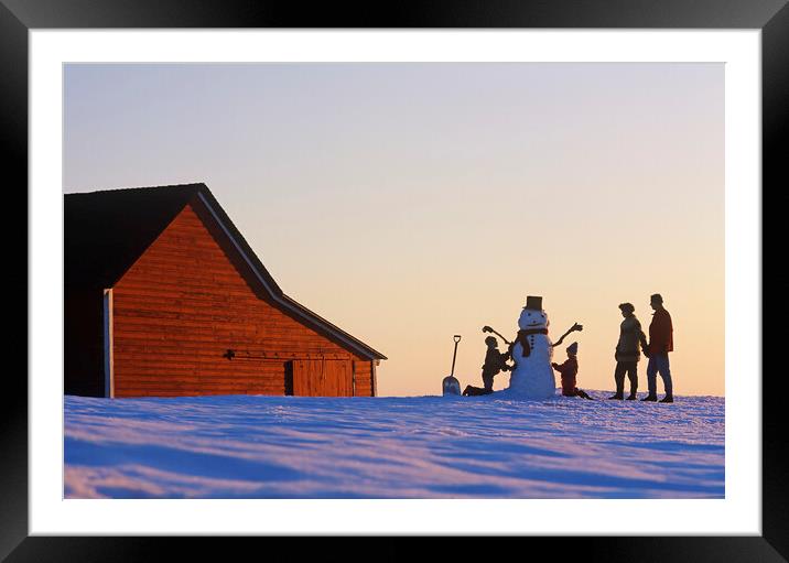 a family make a snowman in front of a red barn Framed Mounted Print by Dave Reede