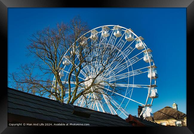 Ferris wheel against a clear blue sky, partially obscured by a rooftop, with bare trees in the background in Lancaster. Framed Print by Man And Life