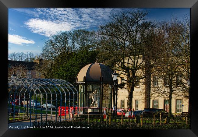 Urban park scene with modern glass pavilion, traditional street lamp, and lush trees under a blue sky with wispy clouds in Harrogate, England. Framed Print by Man And Life