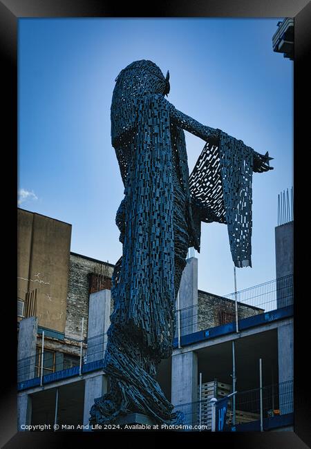 Artistic metal sculpture of a humanoid figure against a clear blue sky, with urban buildings in the background in Leeds, UK. Framed Print by Man And Life