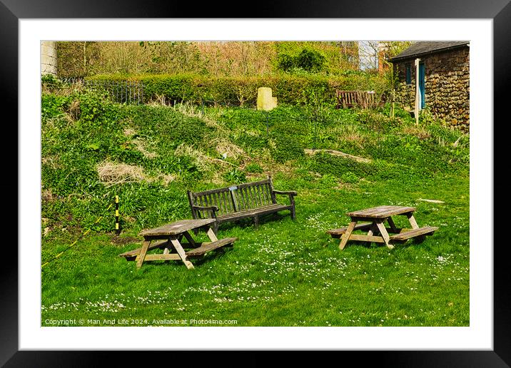 Rustic wooden benches and a table on a lush green grassy hillside, with a stone building and vegetation in the background, depicting a serene outdoor setting in York, North Yorkshire, England. Framed Mounted Print by Man And Life