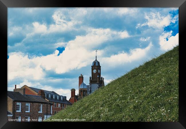 Quaint European town with historic buildings and a clock tower, set against a vibrant blue sky with fluffy clouds, and a lush green hill in the foreground in York, North Yorkshire, England. Framed Print by Man And Life