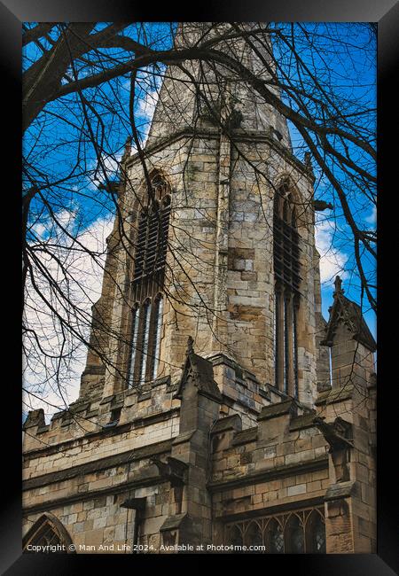 Gothic church tower with intricate stone details, framed by bare tree branches against a blue sky with fluffy clouds in York, North Yorkshire, England. Framed Print by Man And Life
