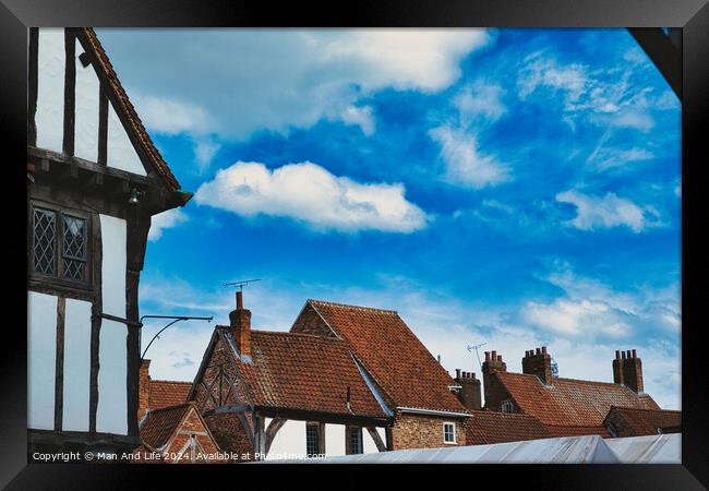 Quaint European village with traditional half-timbered houses and terracotta rooftops under a vibrant blue sky with fluffy clouds in York, North Yorkshire, England. Framed Print by Man And Life