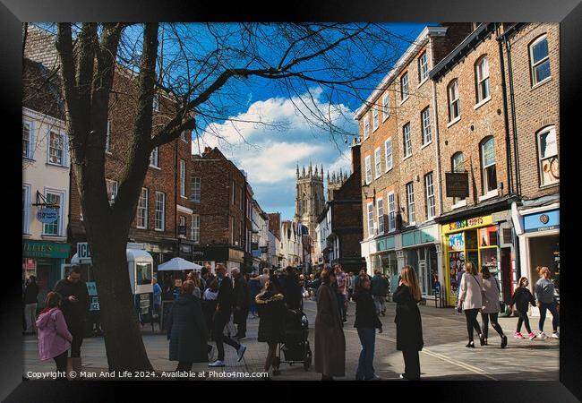 Bustling city street scene with pedestrians, historic buildings, and a cathedral spire under a blue sky with scattered clouds in York, North Yorkshire, England. Framed Print by Man And Life