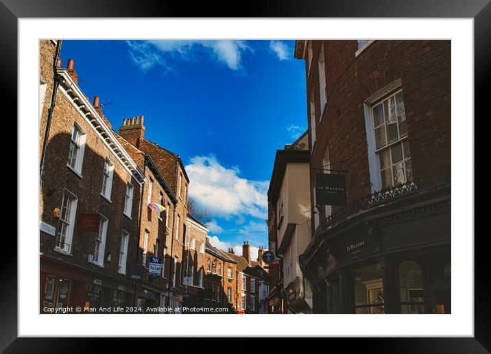 Charming European street scene with historic brick buildings under a clear blue sky with fluffy clouds, showcasing architectural details and local businesses in York, North Yorkshire, England. Framed Mounted Print by Man And Life