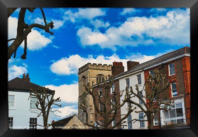 Quaint urban scene with historic stone tower, traditional buildings, and bare tree branches against a vibrant blue sky with fluffy white clouds in York, North Yorkshire, England. Framed Print by Man And Life