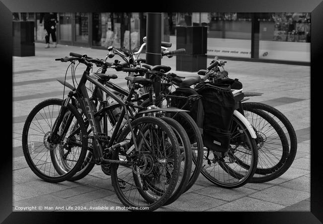 Black and white image of multiple bicycles locked to a bike rack in an urban setting, with a blurred background of a city street Framed Print by Man And Life
