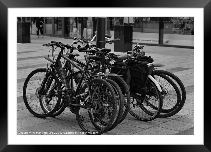 Black and white image of multiple bicycles locked to a bike rack in an urban setting, with a blurred background of a city street Framed Mounted Print by Man And Life