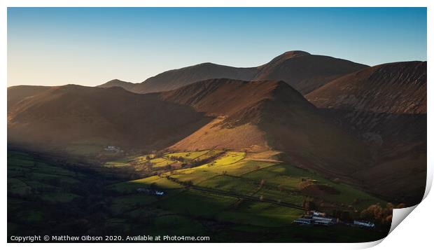 Beautiful Autumn Fall landscape image of sun beams lighting up small area of mountain side in Lake District whilst rest of area is in darkness with Robinson and Dale Head in background Print by Matthew Gibson