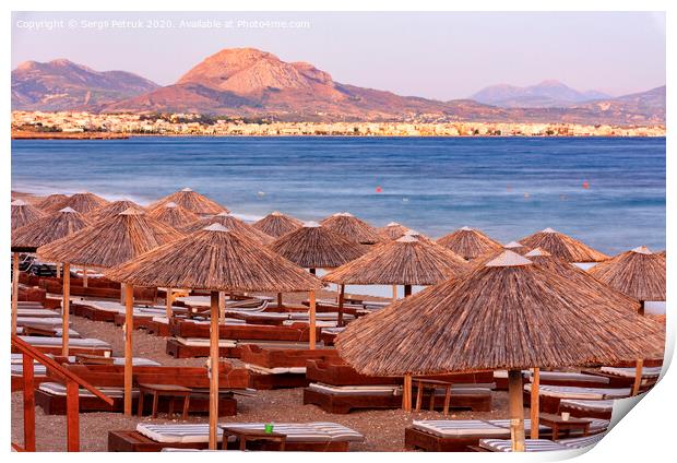 The straw tops of beach umbrellas and wooden deck chairs with mattresses on the deserted beach promenade in the rays of the evening setting sun. Print by Sergii Petruk