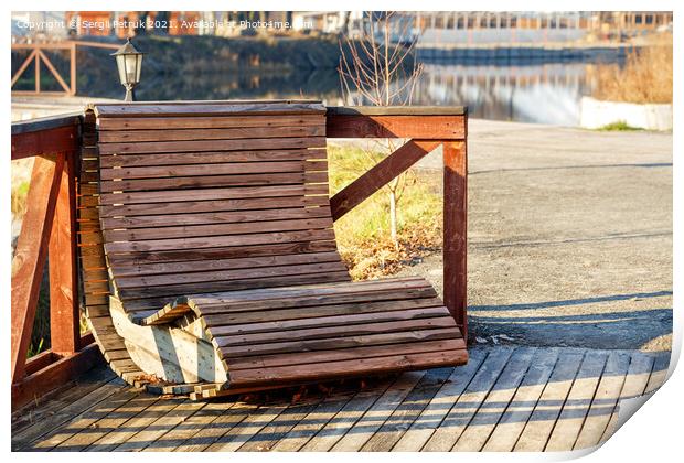 Outdoor furniture, a wooden chair on the river bank is illuminated by the rays of the bright sun against the background of the calm smooth surface of the water in blur. Print by Sergii Petruk