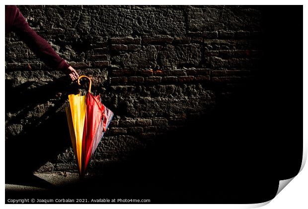 A beautiful colored umbrella rests folded against a brick wall i Print by Joaquin Corbalan