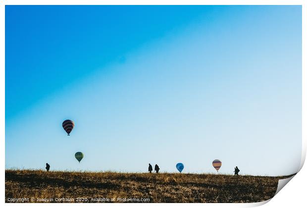 Silhouettes of tourists walking through a meadow while watching hot air balloons flying on the horizon, blue sky background, copy space, added film grain. Print by Joaquin Corbalan