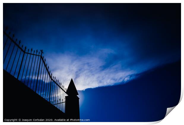 Silhouette against the sun of a high wall and metal fence with an intense blue sky in the background. Print by Joaquin Corbalan