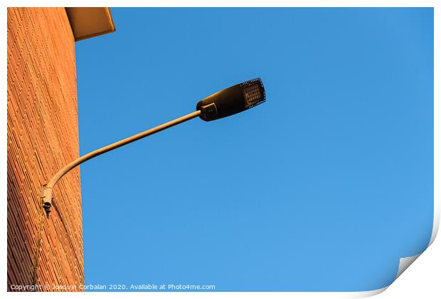 Lamppost on the red wall of a building, by day, with a blue sky background. Print by Joaquin Corbalan