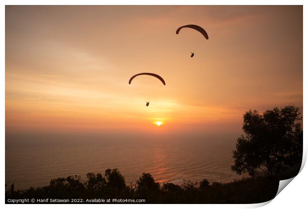 Two paraglider over treetops and ocean at sunset Print by Hanif Setiawan