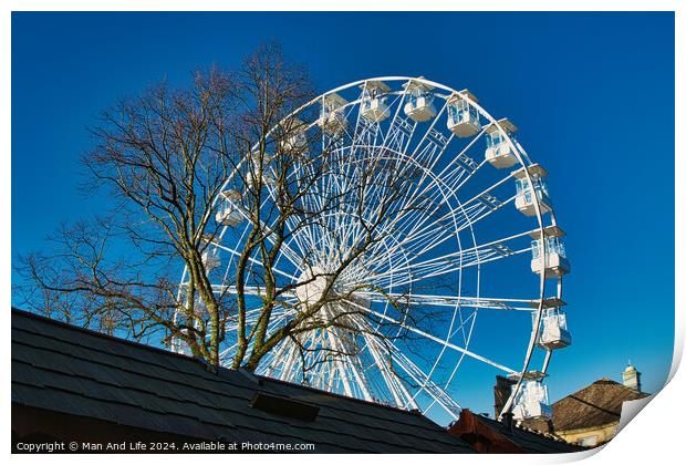 Ferris wheel against a clear blue sky, partially obscured by a rooftop, with bare trees in the background in Lancaster. Print by Man And Life