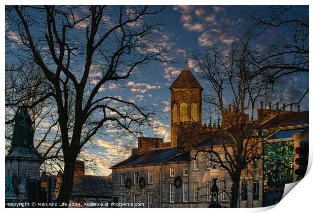Historic stone building with a clock tower at dusk, silhouetted trees, and a vibrant sky in Lancaster. Print by Man And Life