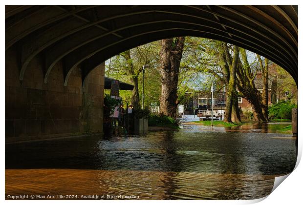 Tranquil river flowing under an arched stone bridge with lush green trees and a hint of urban life in the background, showcasing a blend of nature and city architecture in York, North Yorkshire, England. Print by Man And Life