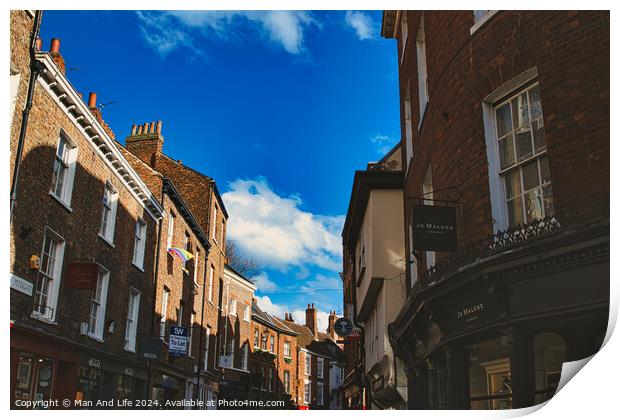 Charming European street scene with historic brick buildings under a clear blue sky with fluffy clouds, showcasing architectural details and local businesses in York, North Yorkshire, England. Print by Man And Life