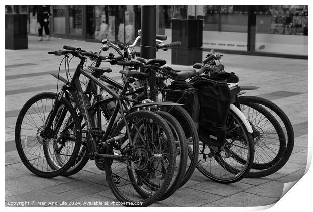 Black and white image of multiple bicycles locked to a bike rack in an urban setting, with a blurred background of a city street Print by Man And Life